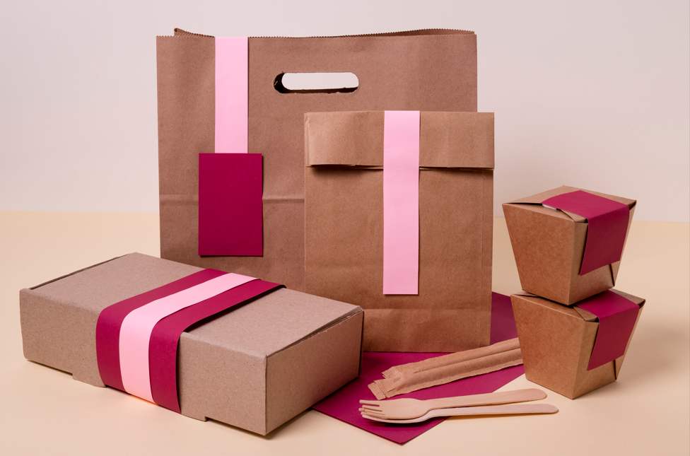 Eco-friendly packaging set including paper bags, boxes, and containers with pink & burgundy accents, styled with wooden cutlery