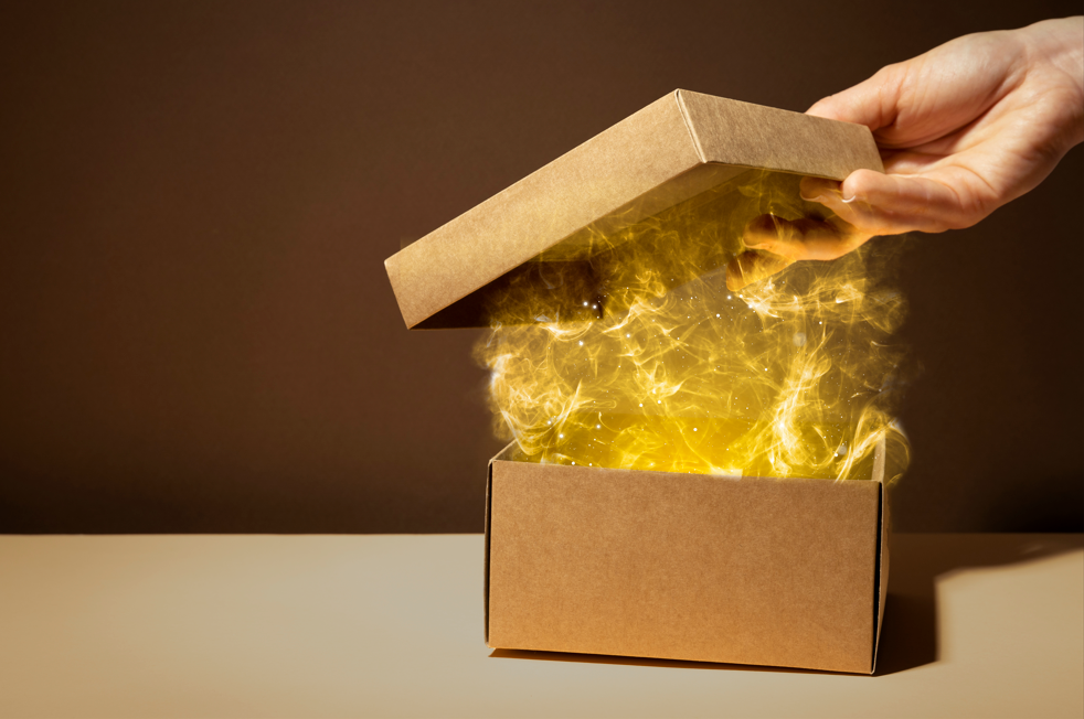 Hand lifting the lid of a cardboard box with glowing golden light and spark-like energy rising out, symbolizing creativity, innovation, or mystery