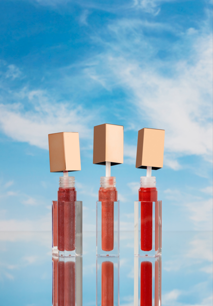 Three tubes of lip gloss in different shades with gold caps, upright on reflective surface against a bright blue sky